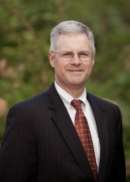 white man with gray hair and glasses in black suit with red tie